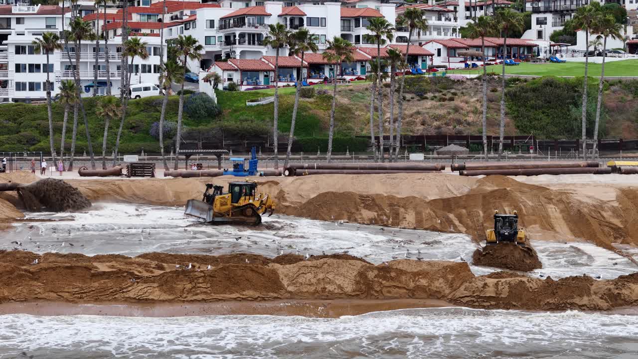 las excavadoras trabajan en el reemplazo de arena en una playa con casas costeras y palmeras en el fondo