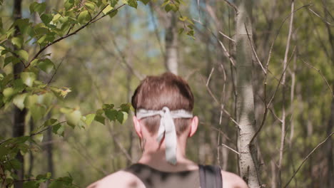 back view of youthful adventurer wearing snug headband carrying bag carefully navigating through dense forest undergrowth while pushing aside branches and foliage under sunlight on woodland trail