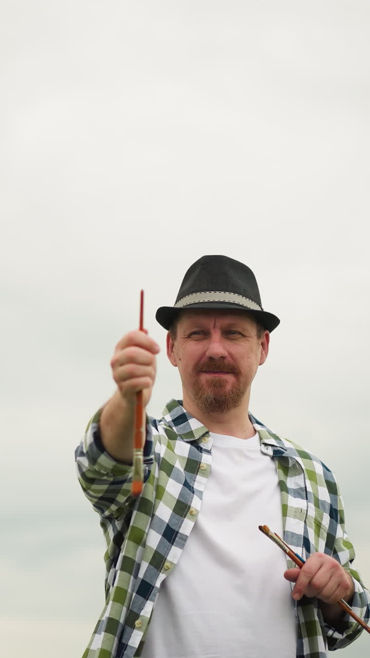 vista frontal de un artista con un sombrero y una camisa a cuadros, sosteniendo un pincel y apuntándolo hacia adelante para determinar un ángulo. el artista está enfocado, composición perfecta en un entorno al aire libre
