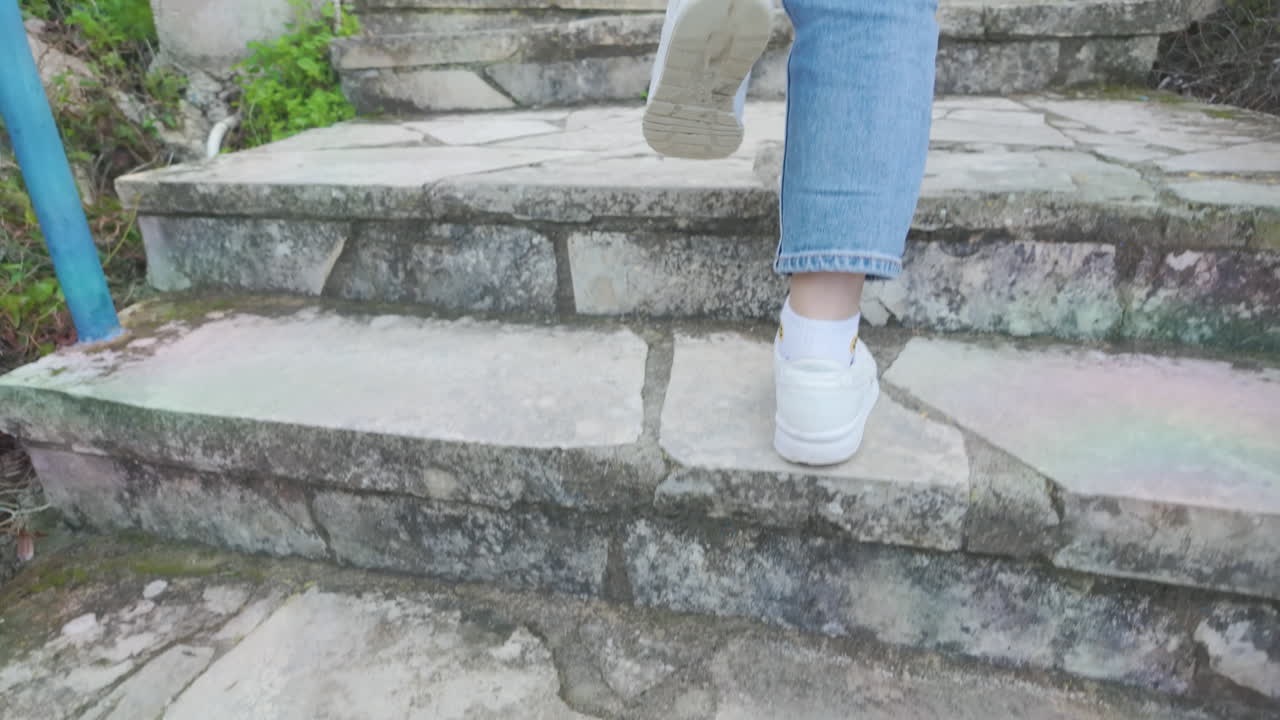 primer plano de la mitad inferior de una persona, con pantalones vaqueros y zapatillas blancas, subiendo escalones de piedra
