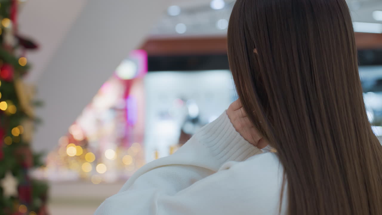 Close-up of woman leaning on railing adorned with festive lights in shopping mall, slight blur in background with people passing by, thoughtful pose with hand on chin