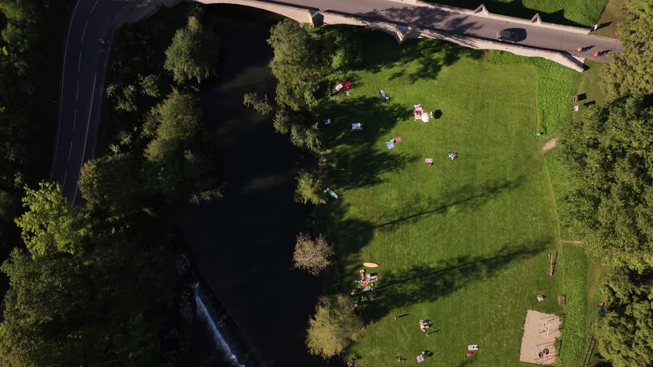 Top-down shot of a dam next to a riverbank with people and children swimming, playing games and sunbathing on a lawn