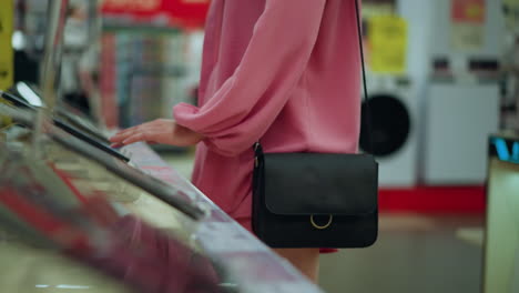 body view of lady in pink dress with black handbag interacting with a tablet on display stand in bright tech store, screen is operated while device sits on sleek stand, blurred person in background