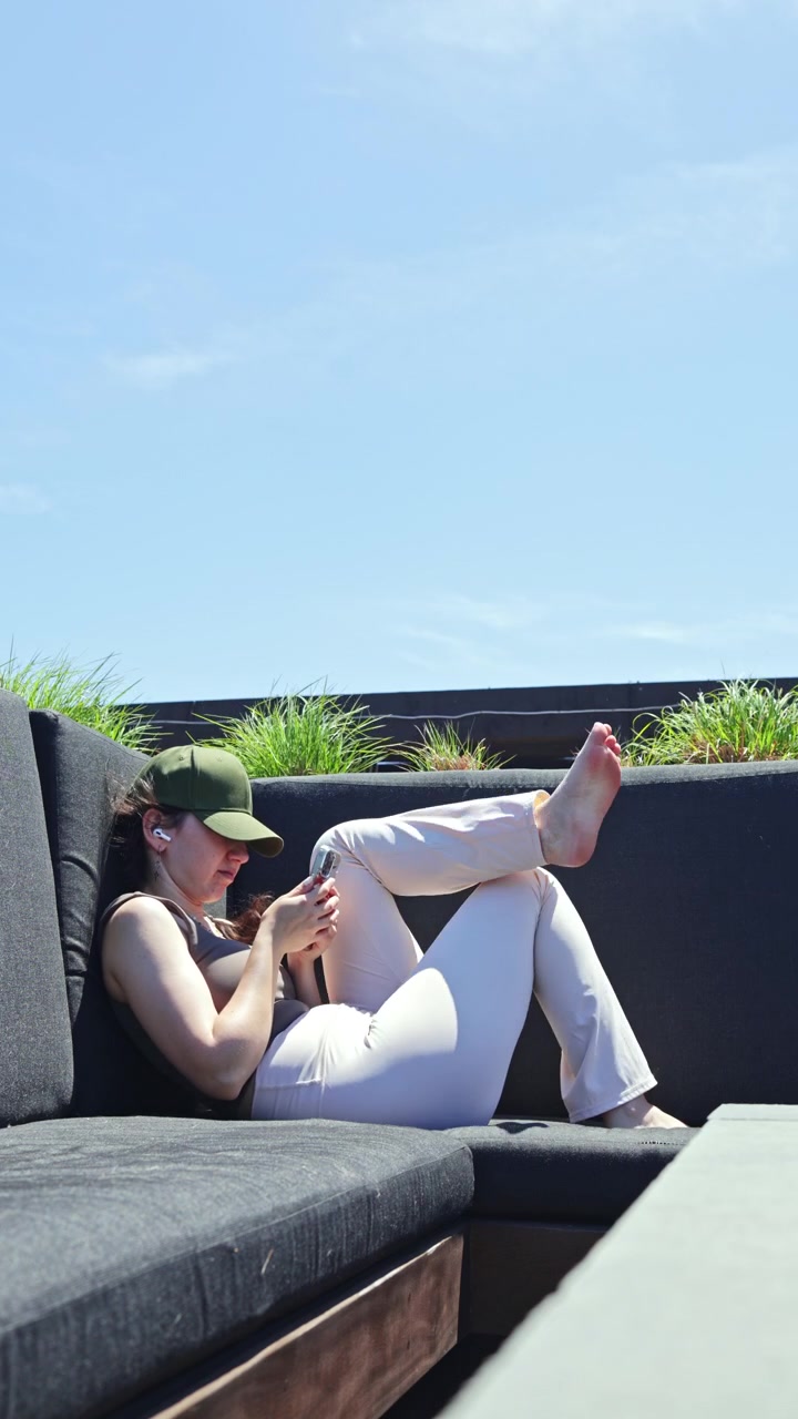 Woman sitting on a couch using a smartphone in an outdoor space during clear weather surrounded by greenery