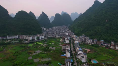 Aerial-hyperlapse-captures-cars-driving-from-farm-fields-to-Yangshuo-city,-nestled-amidst-green-mountains-at-sunrise