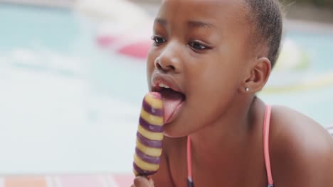 una chica afroamericana feliz sonriendo y comiendo helado junto a la piscina, en cámara lenta, sin alteración.