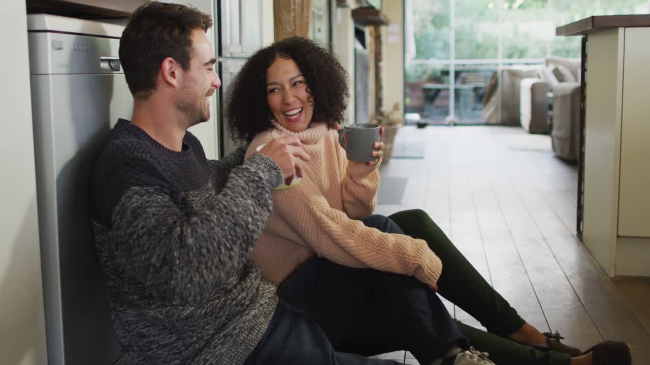 Happy diverse couple talking and drinking coffee sitting on floor in kitchen