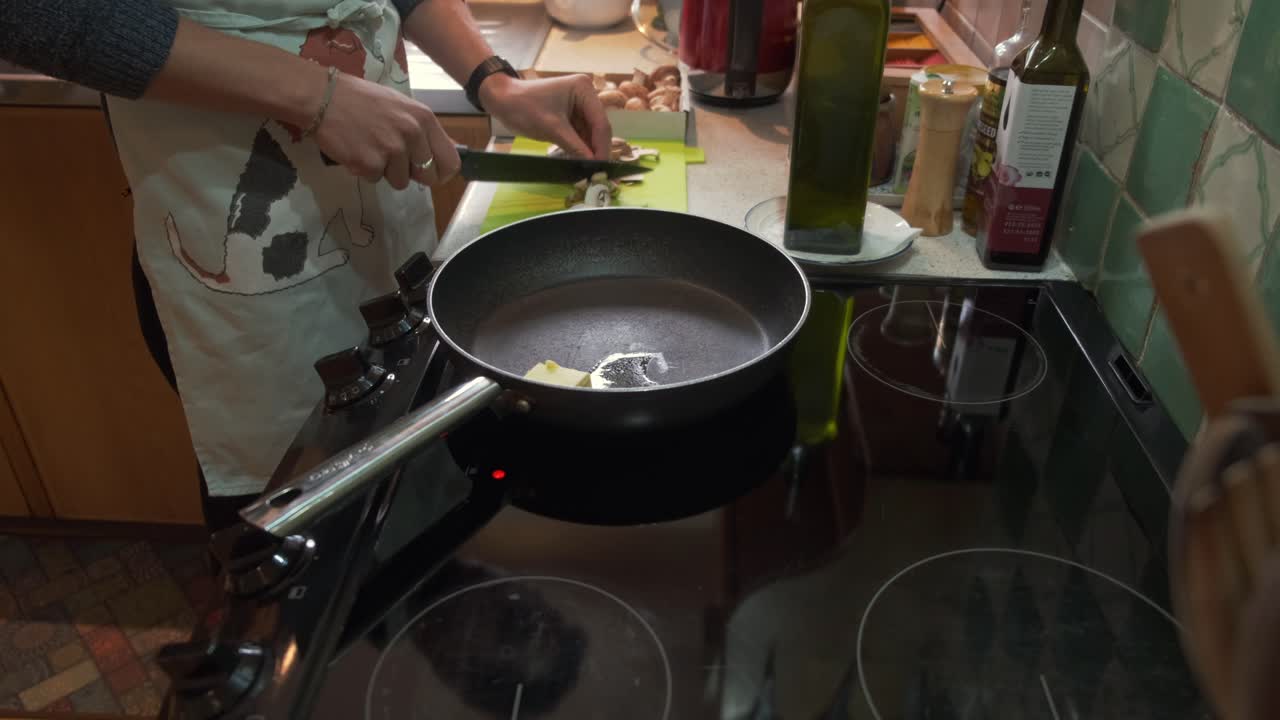 A white male cook in an apron melts butter in a frying pan on the stove while slicing mushrooms with a knife over a tray beside the pan in his cozy home kitchen.