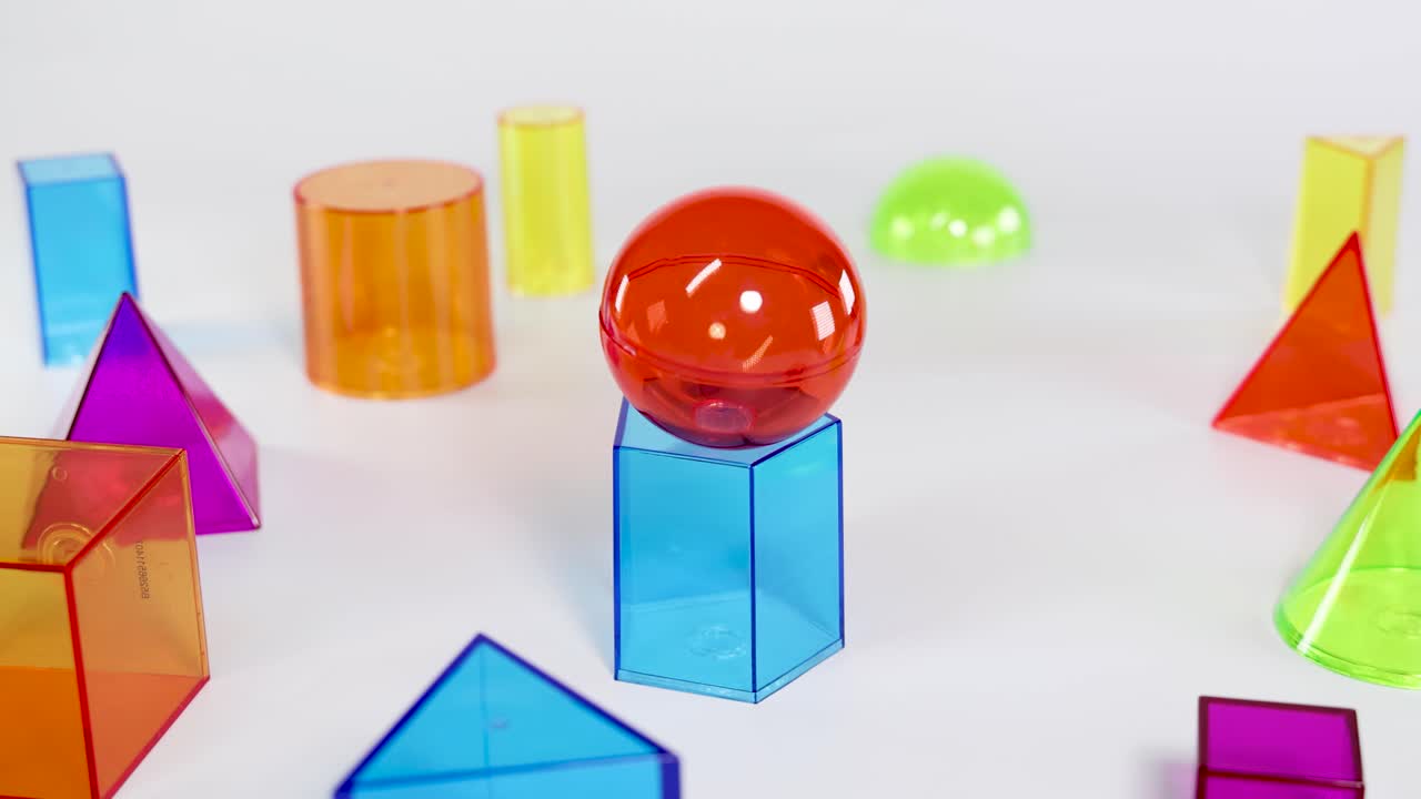 Uma mão equilibra cuidadosamente uma esfera vermelha em cima de um prisma hexagonal azul sobre um plano de fundo branco