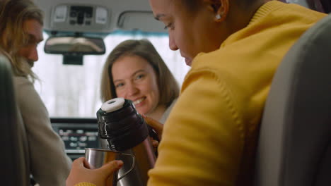 rear view of man sitting in the back seat of a car pouring tea from a thermos into a steel mug and serving it to woman