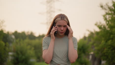 young woman appears exhausted and frustrated during phone call, covering face with her left hand while talking, set against a blurred outdoor background of greenery and distant buildings