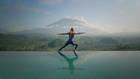 mujer lanzándose a la pose de yoga guerrera en el borde de la piscina infinita con el monte agung al amanecer