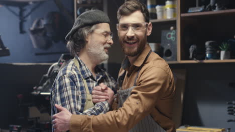 portrait shot of two cheerful father and son in goggles hugging and shaking hands in workshop