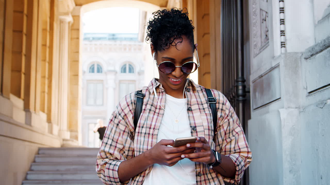 Young adult woman wearing a plaid shirt standing in front of a historical building entrance using her smartphone
