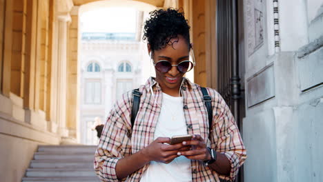 Mujer-Adulta-Joven-Con-Una-Camisa-A-Cuadros-Parada-Frente-A-La-Entrada-De-Un-Edificio-Histórico-Usando-Su-Teléfono-Inteligente