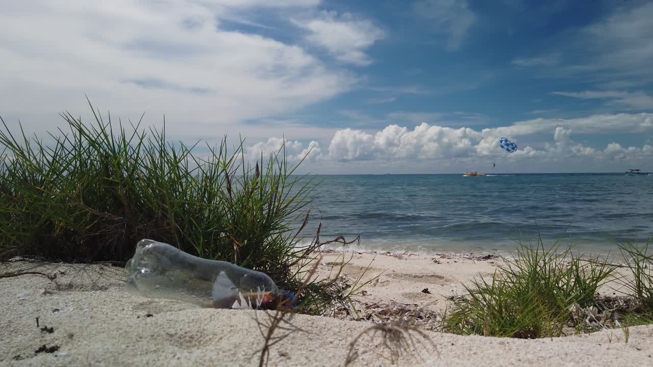 낮은 각도, 느린 동작으로 볼 때, 깨한 플라스틱 소다 병은 평온한 색 물과 놀라운 파란 하늘을 가진 아름다운  모래 해변에 버려집니다.