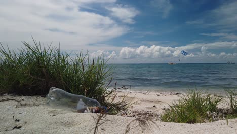 low angle, slow motion view of a clear plastic soda bottle discarded on a beautiful white sand beach with calm turquoise water and stunning blue sky