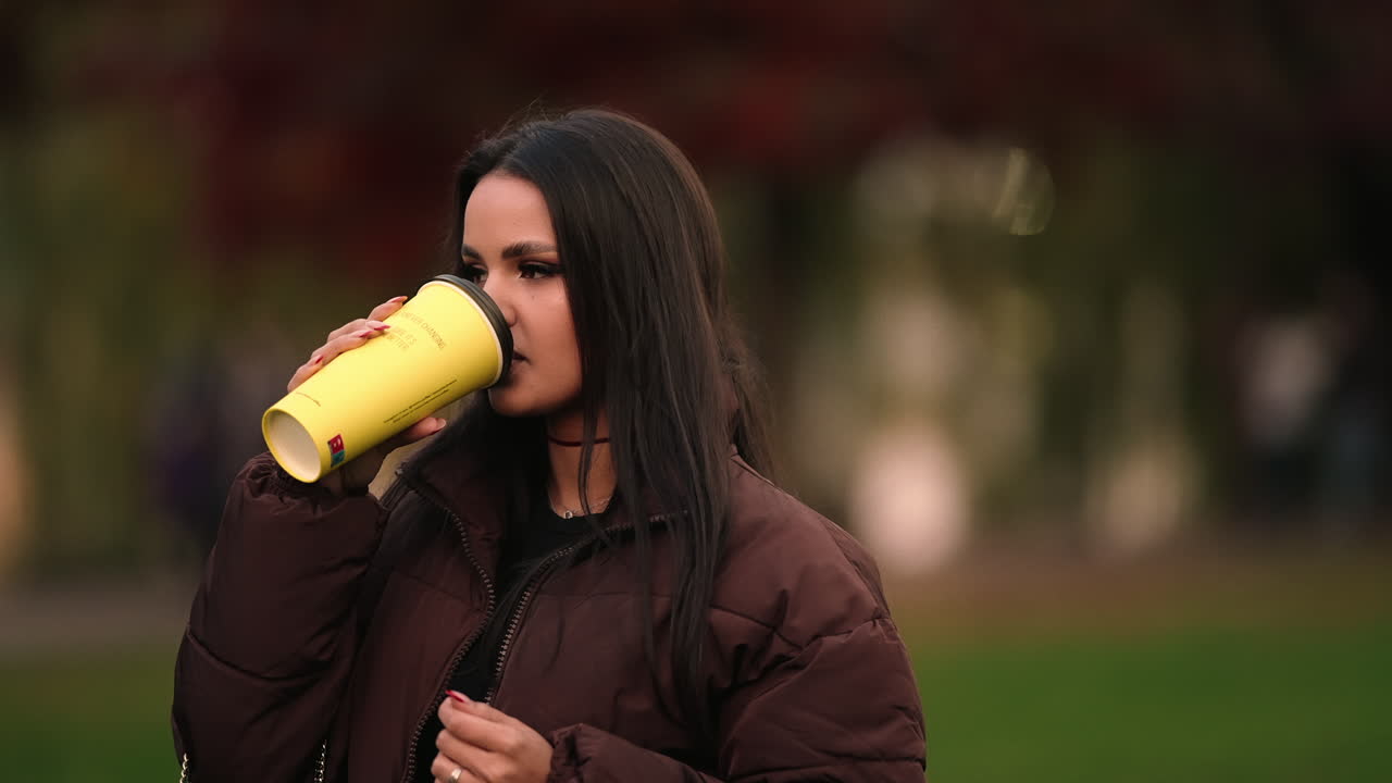 Ritratto di bellissima giovane ragazza modella mora che tiene e beve una tazza di caffè in un parco all'aperto, sfondo sfocato