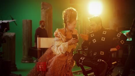 beautiful smiling actress wearing renaissance dress and actor wearing motion capture suit sitting on chairs share social media posts via smartphone. on film studio period costume drama film set