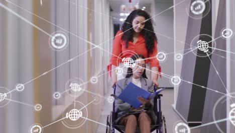 Animation-of-connected-graph-and-globe-icons,-diverse-woman-assisting-coworker-sitting-on-wheelchair