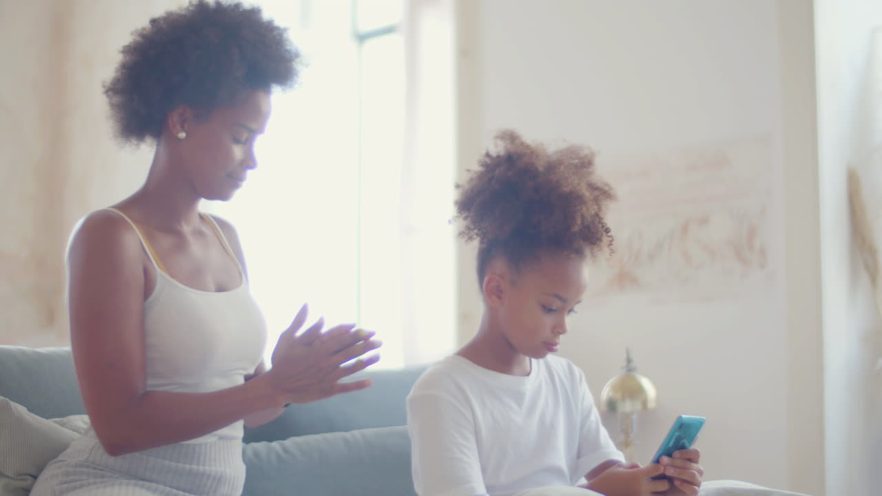 Mãe afro-americana penteando o cabelo da filha enquanto a menina ouve música no celular e dança.