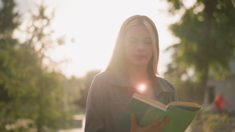 lady in grey clothing walking on grassy field while reading a book as sunlight creates bright flashes around her, soft blur of trees and a person cycling in the background