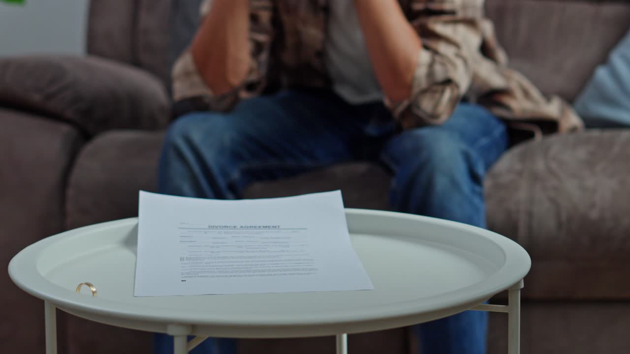 Wife hand dropping wedding band falling onto legal divorce papers while distraught husband sits