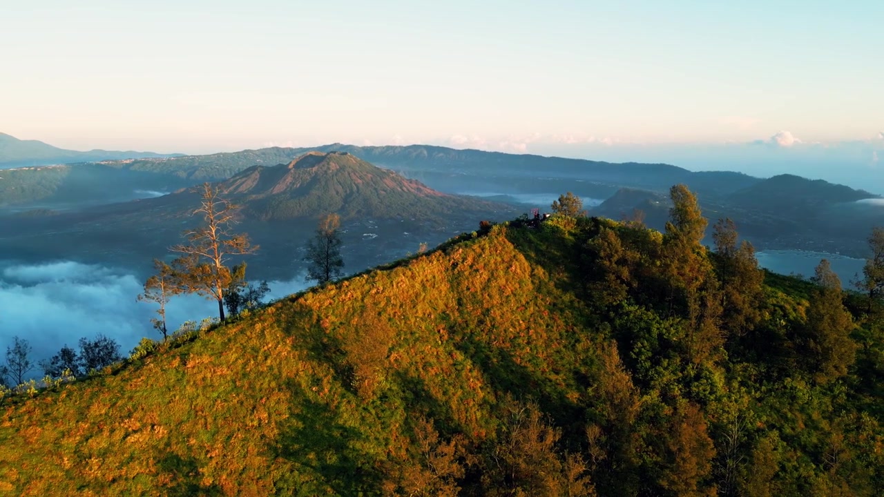 Gunung berapi Batur dan danau kawah dengan lereng gunung hijau dalam cahaya pagi keemasan melintasi medan dataran tinggi di Bali dengan lautan awan, rekaman drone udara yang mulus.