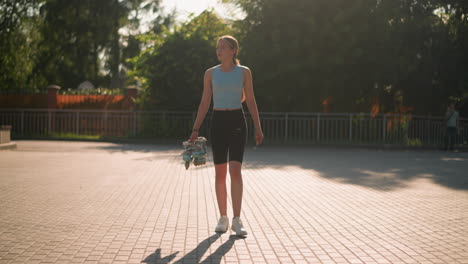 lady wearing athletic wear walking on interlocked path holding roller skates under warm sunlight with greenery in background, featuring soft shadows and another person in blurred distance