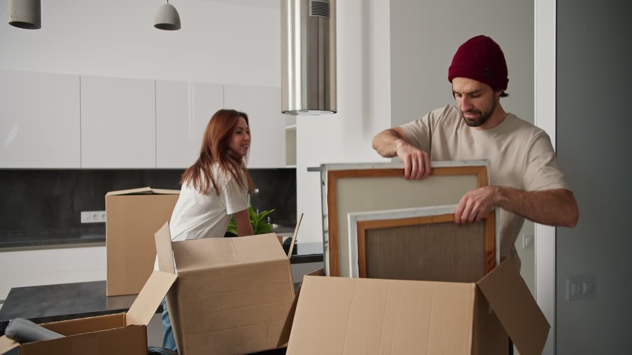 hombre feliz con paja en una gorra roja le da a su novia morena en una camiseta blanca cosas de cajas grandes durante su mudanza en un apartamento moderno