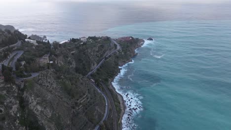 An aerial shot of a mountain slope next to the sea, with a winding road running along the hillside. The water below is a vibrant azure.