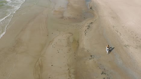 Aerial-view-with-a-young-longhaired-girl-riding-a-bike-on-the-sandy-beach,-sunny-day,-white-sand-beach,-active-lifestyle-concept,-wide-ascending-birdseye-drone-shot-moving-forward