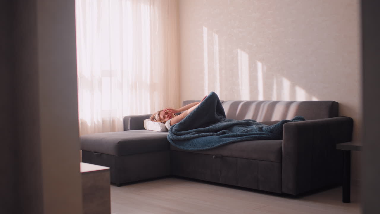 Young girl stretches after sleep on couch covered with blanket, relaxing in warm morning sunlight streaming through curtains, cozy home atmosphere reflecting comfort