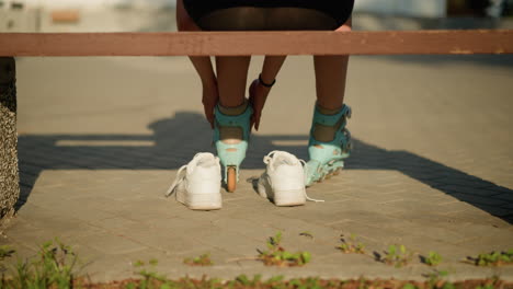 back view of individual sitting on bench adjusting straps on cyan roller skates on left leg while warm sunlight casts shadows on pavement, white sneakers are resting behind, greenery peeks from edges