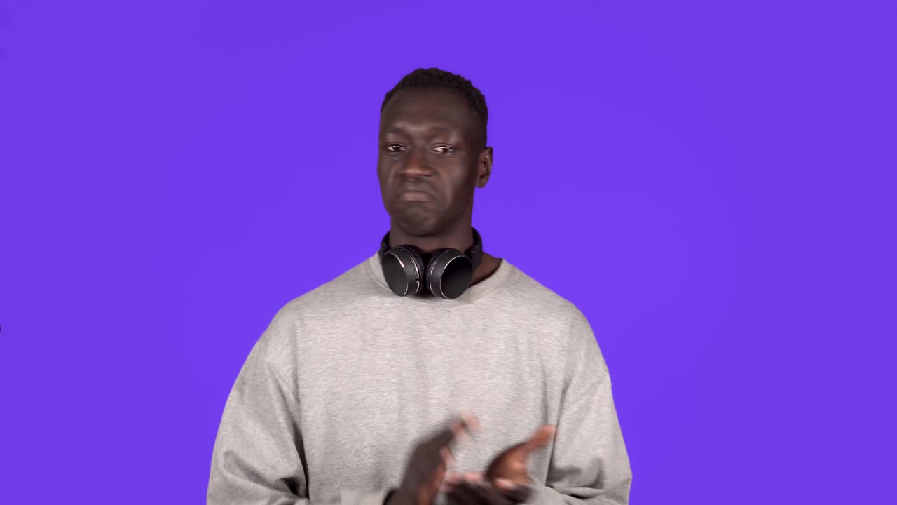 Clapping young african man, applauding in respect to someone. Isolated on blue background. Wearing white sweater and headphones on neck