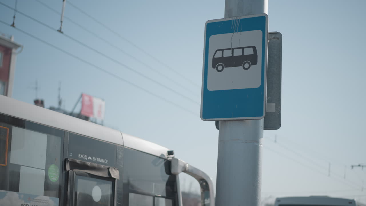 close view shows bus stop sign on pole as bus passes along city street, overhead wires cut blue winter sky, buildings blur behind, urban transit moment with motion, public transport symbol foreground