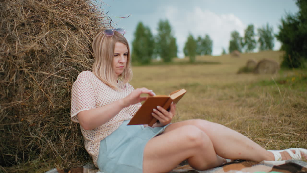 Young woman sitting on blanket, resting against hay bale, flipping book pages thoughtfully, green notebook beside her on grass, blurred rural background with trees and hay bales in distance