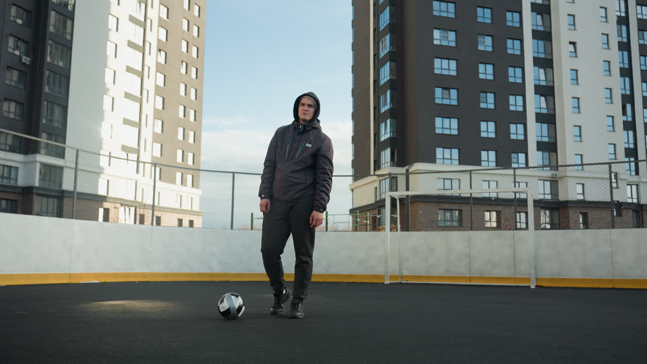 후디를 입은 선수가 스포츠 경기장에서 자신감 있게 서 있고, 앞에 공을 놓고 다리를 돌리고, 도시 배경에는 은 하늘 아래 높은 현대 사무실 건물이 있습니다.