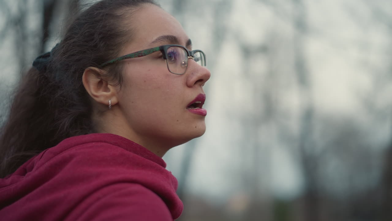 Young Woman In Red Hoodie Looking Upward In Winter Woods, Profile Closeup, Glasses And Ponytail, Soft Dusk Light, Blurred Bare Trees, Pensive Expression, Shallow Depth, Cinematic Mood