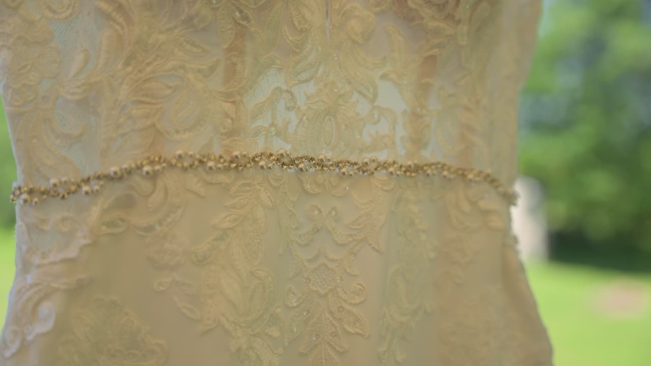 Un gros plan magnifique mettant en valeur la ceinture scintillante en strass et la délicate dentelle florale d'une robe de mariée, avec un jardin d'été flou visible dans la lumière naturelle de l'arrière-plan.