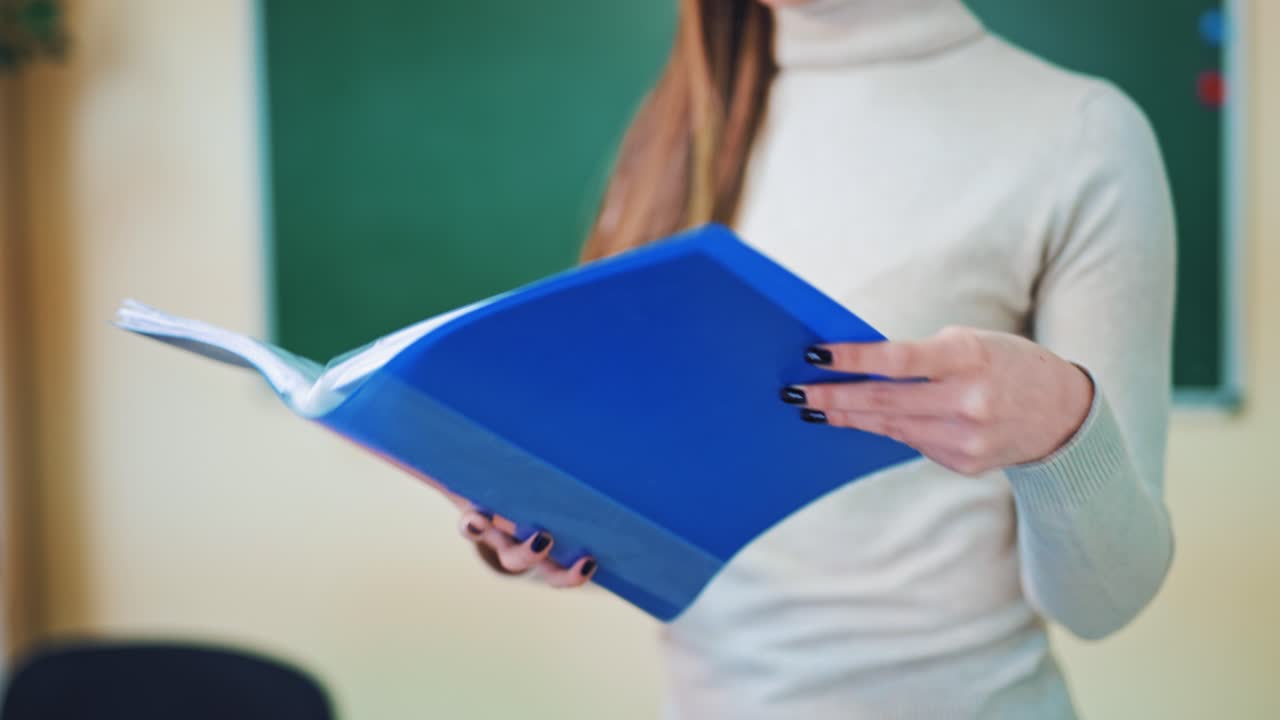 Blue folder in woman's hands in class. Woman turns pages with printed papers in files on educational center background. Close-up.
