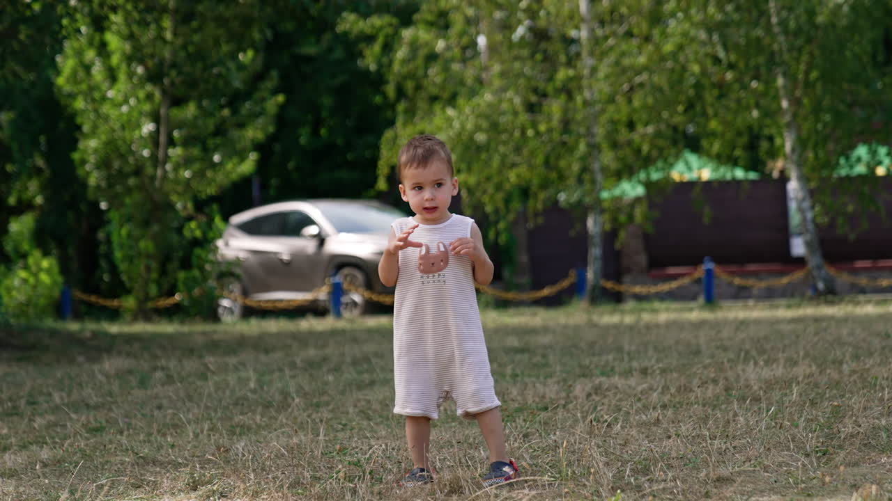Funny baby boy in a romper turns standing on the grass. Little Caucasian child having good time in the summer park.