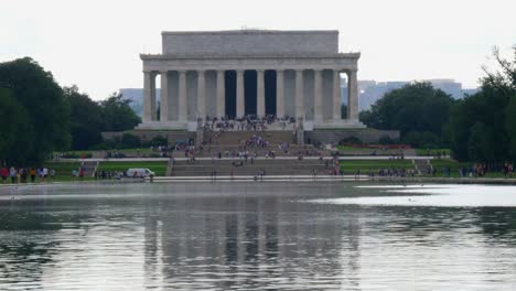 Inclinación-Lenta-Hacia-Arriba-Desde-El-Agua-Hasta-Los-Escalones-Delanteros-Del-Monumento-A-Lincoln-Tomados-Del-Estanque-De-Reflexión-Con-Personas-En-Primer-Plano