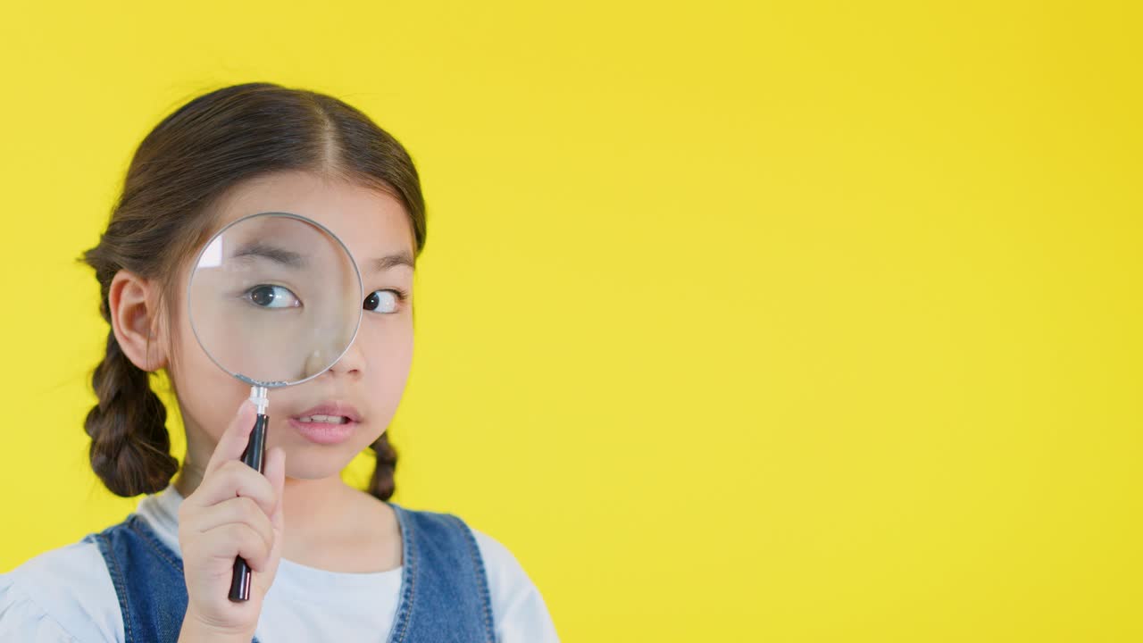 Young girl distorts her face with magnifying glass against bright yellow background, cheerful and playful