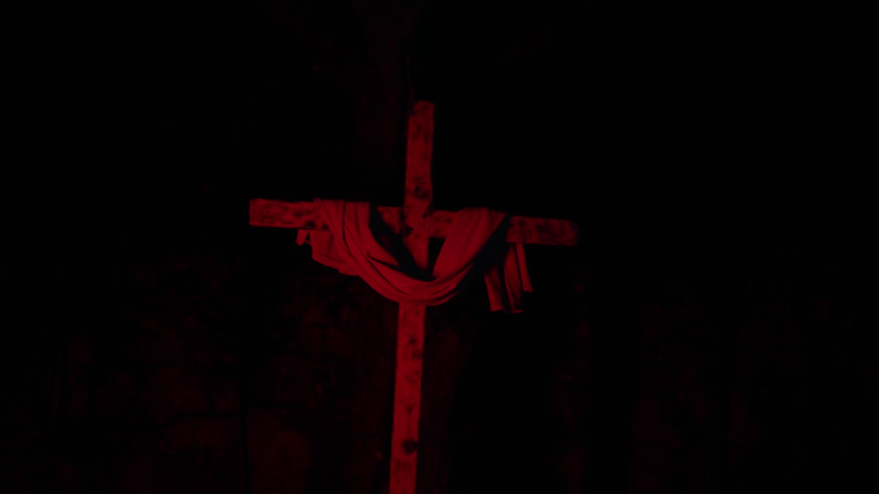 Cruz de madera con tela drapeada sobre ella, iluminada con luz roja intensa sobre un fondo negro.