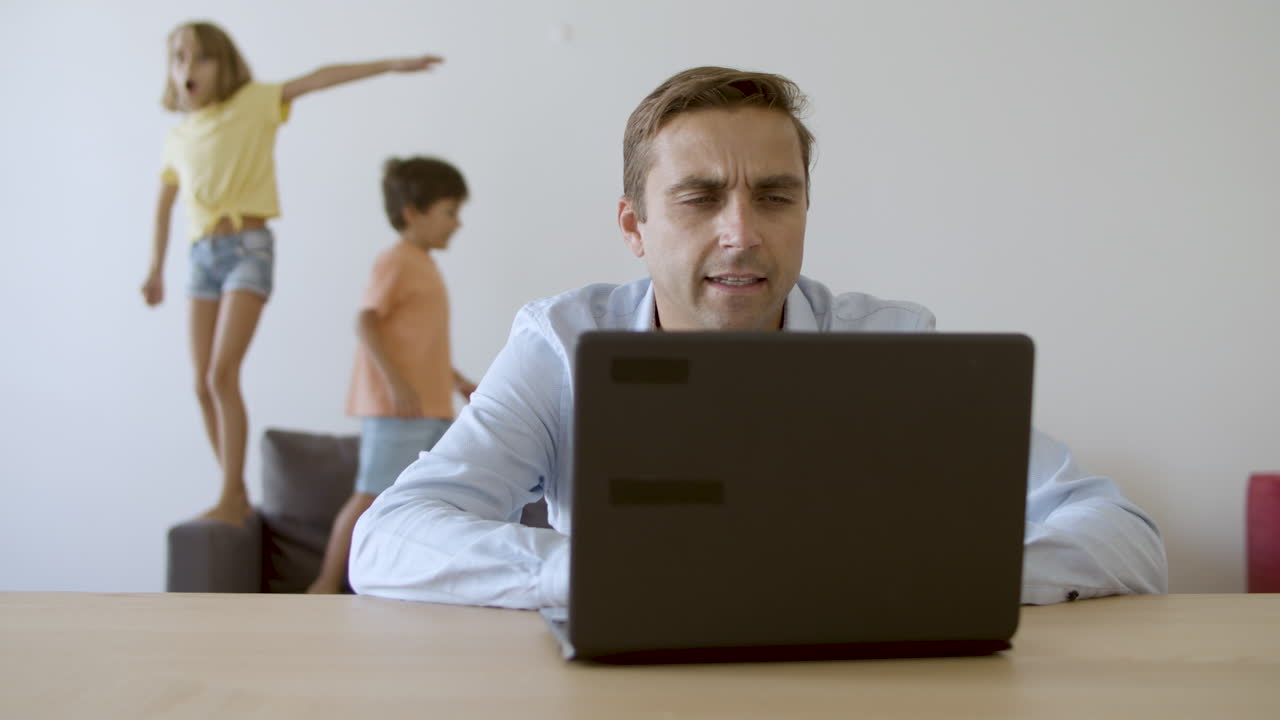 padre serio trabajando a través de la computadora portátil y los niños saltando en el fondo