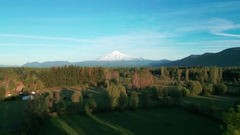 Forward drone approach to snow-capped Villarrica Volcano over dense forest and green fields, bathed in golden light under clear blue skies