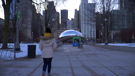 one woman in a coat walks towards city buildings and the cloudgate landmark in chicago, illinois on a cold day.