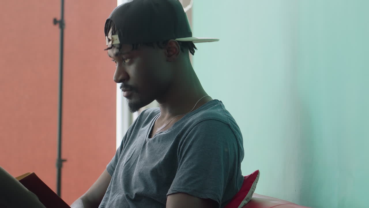 Vista lateral de un hombre joven con camiseta informal y gorra hacia atrás levantando la cabeza de un libro con expresión concentrada, sentado en el interior contra una pared color verde azulado y un fondo cálido