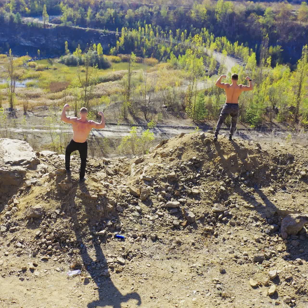 Backside view of two sportsmen on the rocky hill. Shirtless athletes showing their muscular body on the nature background in summer. Aerial view.